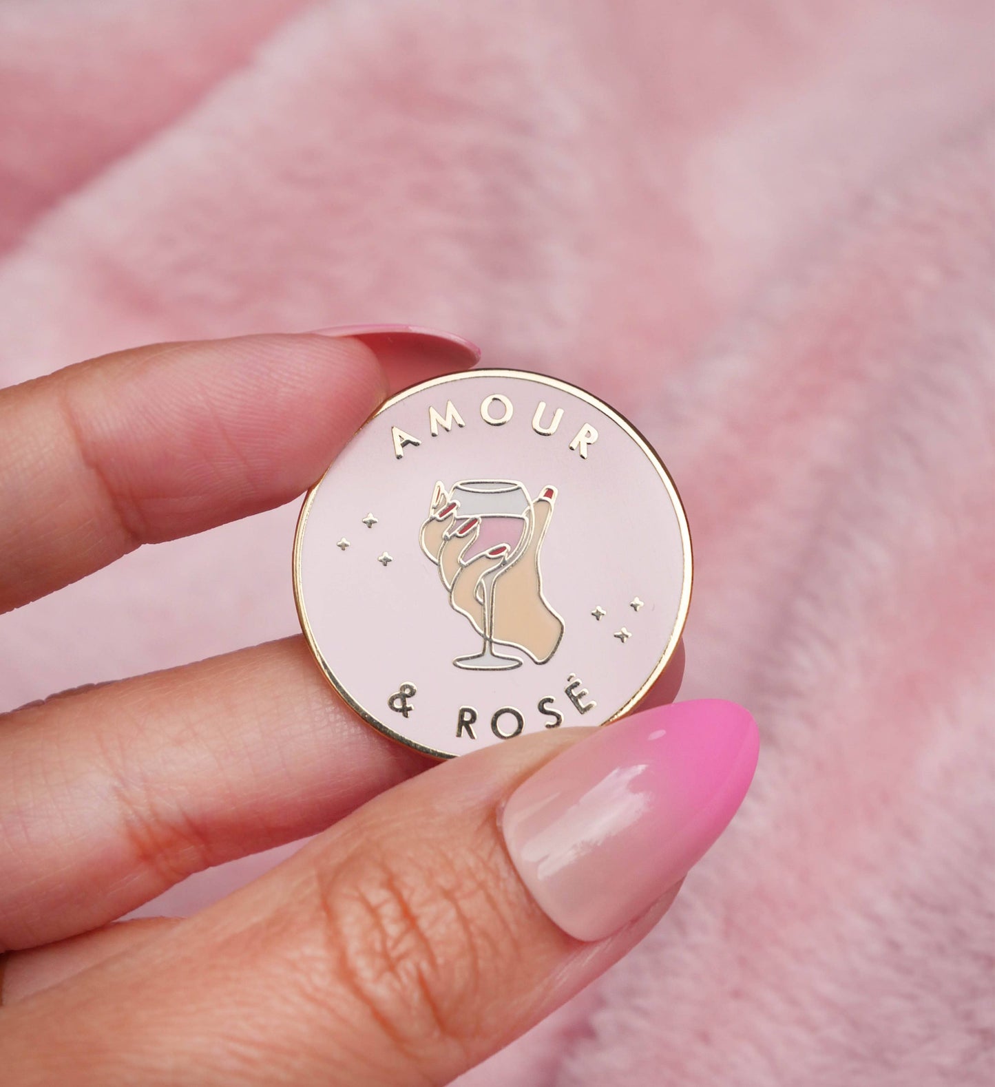 Pin’s doré Amour & Rosé tenu entre deux doigts sur fond rose.