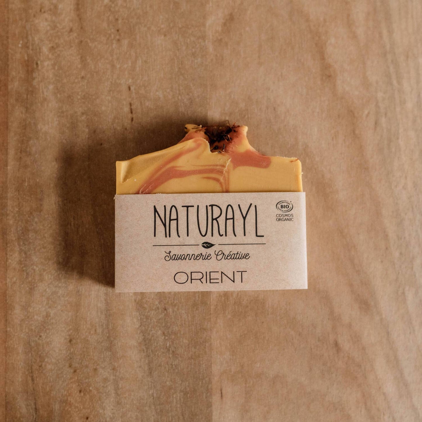« Savon artisanal Naturayl Orient, présenté avec son emballage kraft sur un fond en bois, marbrures jaunes et orangées visibles sur le dessus. 