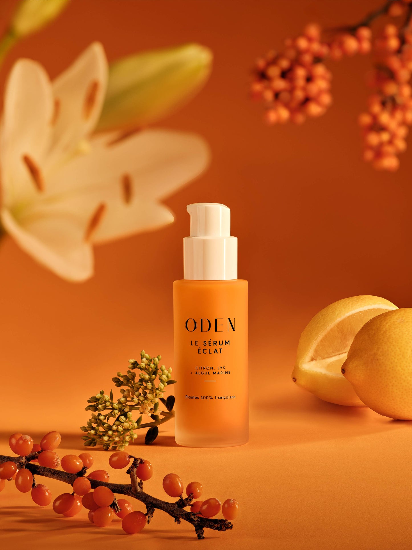 Flacon du Sérum Éclat et Anti-Taches Oden, mis en scène sur fond orangé avec fleurs et agrumes. Soin naturel illuminateur à base de bio-rétinol et d’extraits antioxydants, disponible sur Anahata Concept Store.