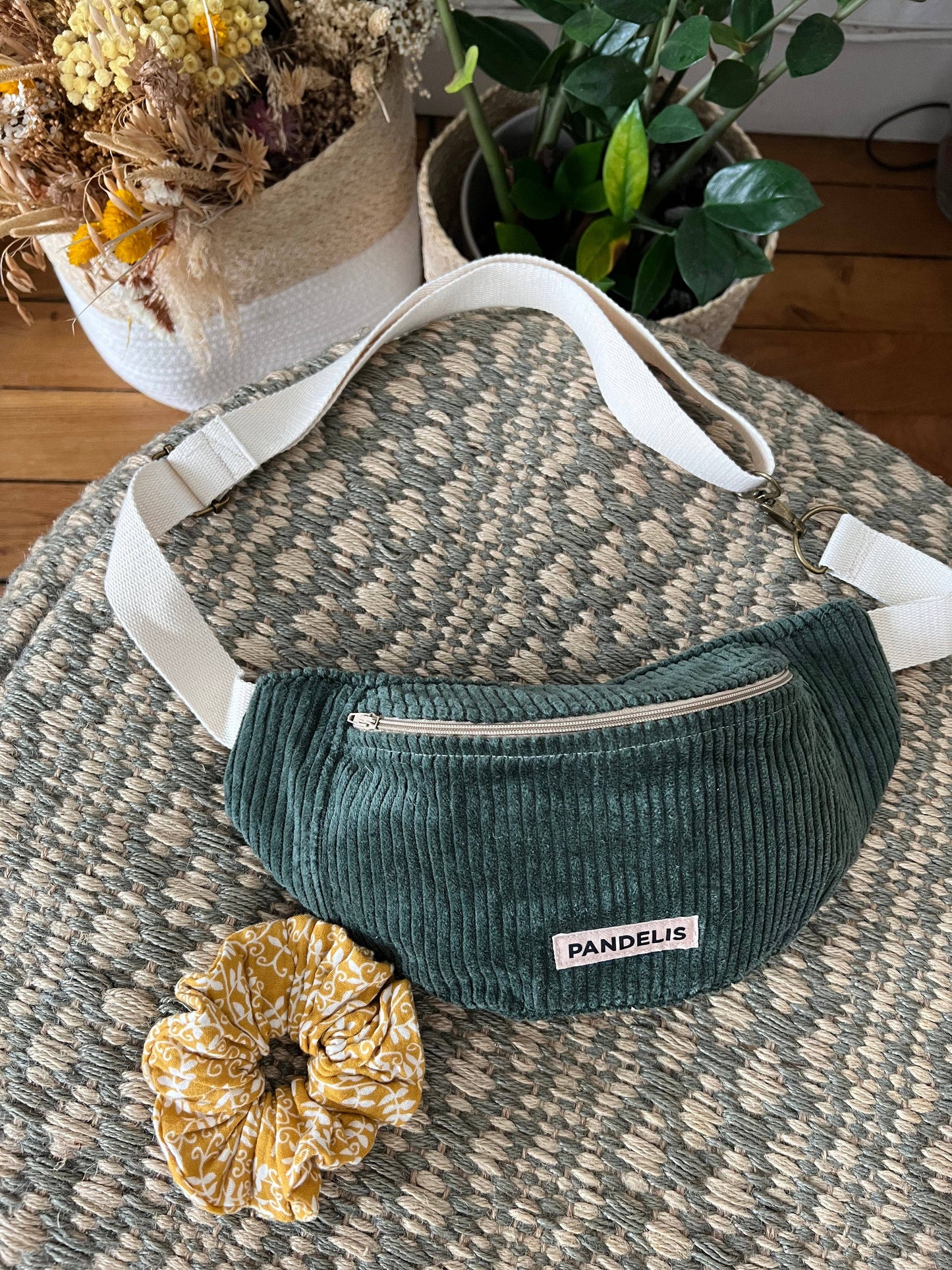 Banane Pandelis vert sauge en velours côtelé, présentée sur une table à motifs avec des éléments déco naturels autour.