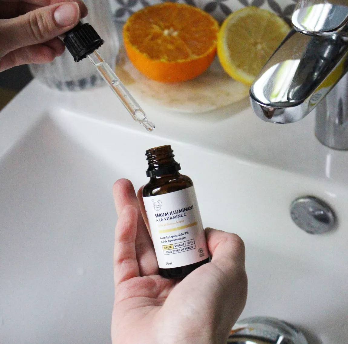 Main tenant la pipette du Sérum illuminant Clémence & Vivien au-dessus d’un lavabo, avec une orange coupée en arrière-plan.