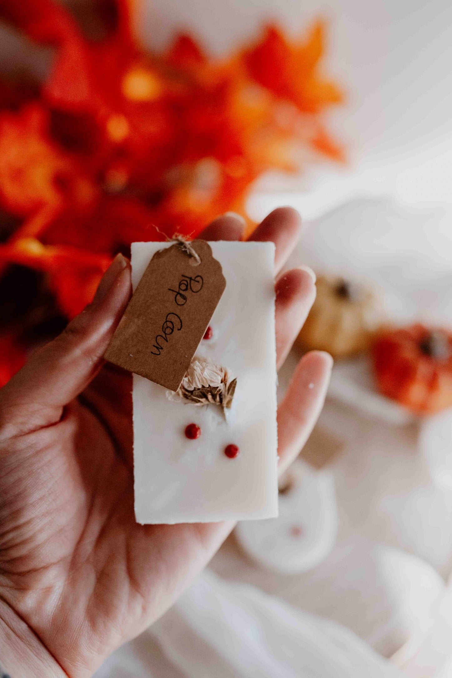 Main tenant une tablette de cire parfumée Pop Corn décorée de fleurs séchées, sur un fond d’ambiance automnale.