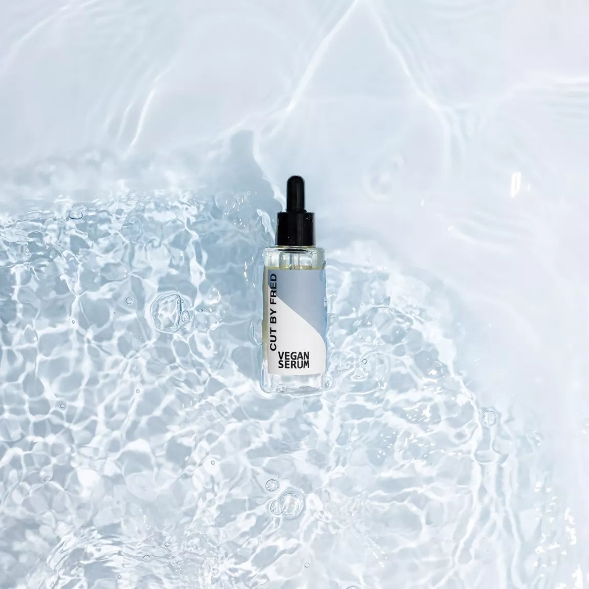 Vegan Serum Cut By Fred, flacon avec pipette posé dans une eau claire, sérum capillaire nourrissant et hydratant.