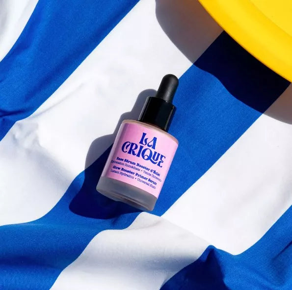Flacon rose du Sérum de base éclat La Crique posé sur un tissu rayé bleu et blanc avec une touche de jaune en arrière-plan.