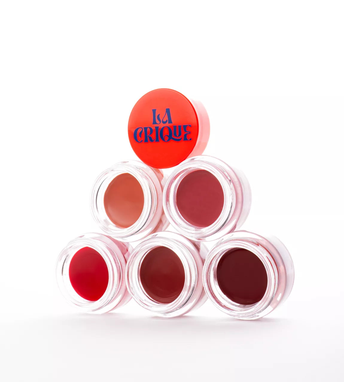 Pyramide de six baumes lèvres et joues hydratants teintés La Crique, présentés dans des pots en verre transparent avec un capot orange vif, logo bleu sur le pot du dessus.