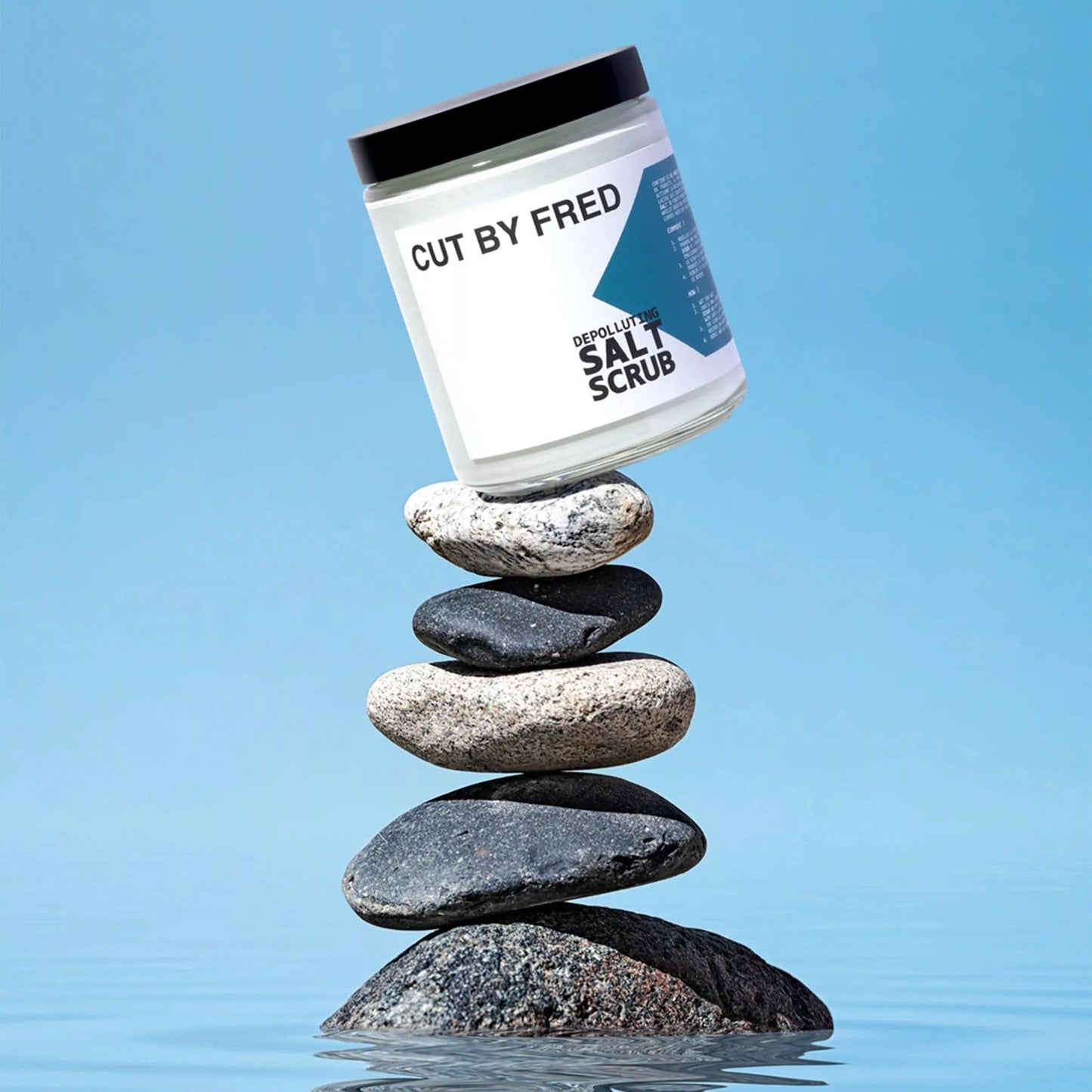 Depolluting Salt Scrub Cut By Fred, pot posé sur une pile de galets, symbole de pureté et d’équilibre.