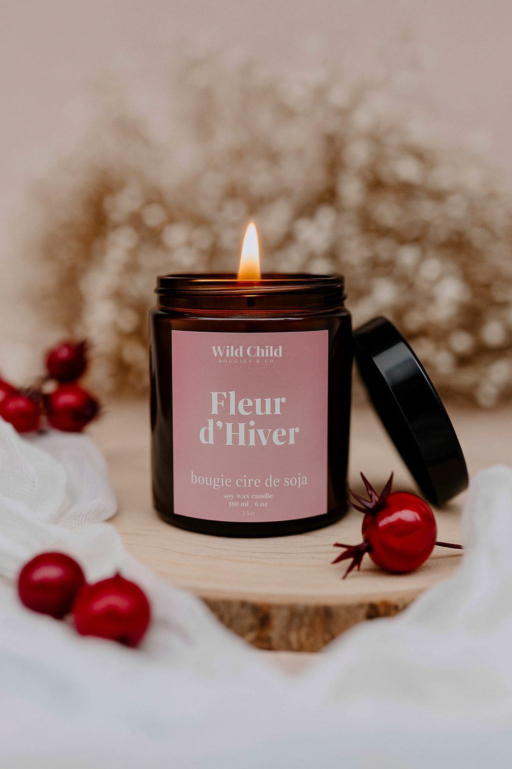Bougie Fleur d’Hiver allumée, pot ambré posé sur un rondin de bois, décor hivernal doux avec baies rouges et gypsophile.