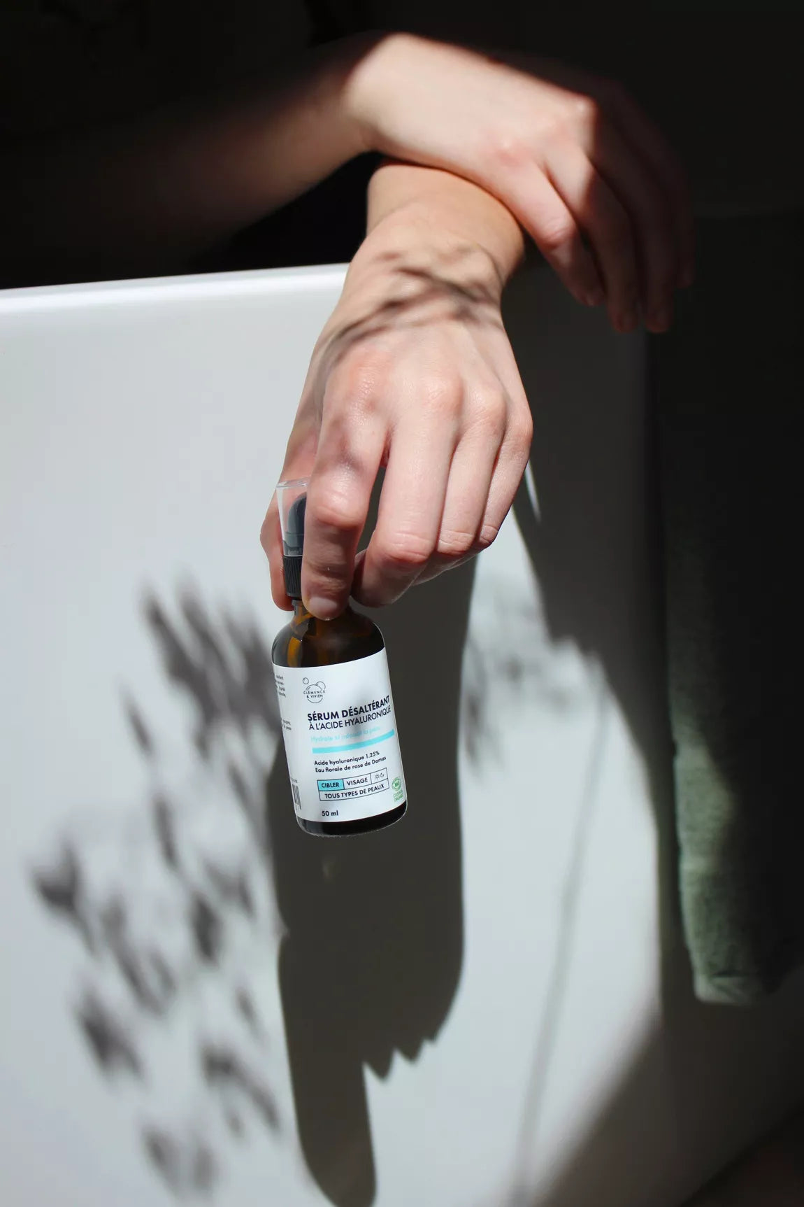 Main posée sur le rebord d’une baignoire tenant le flacon de Sérum désaltérant Clémence & Vivien, avec des jeux d’ombre végétale en arrière-plan.