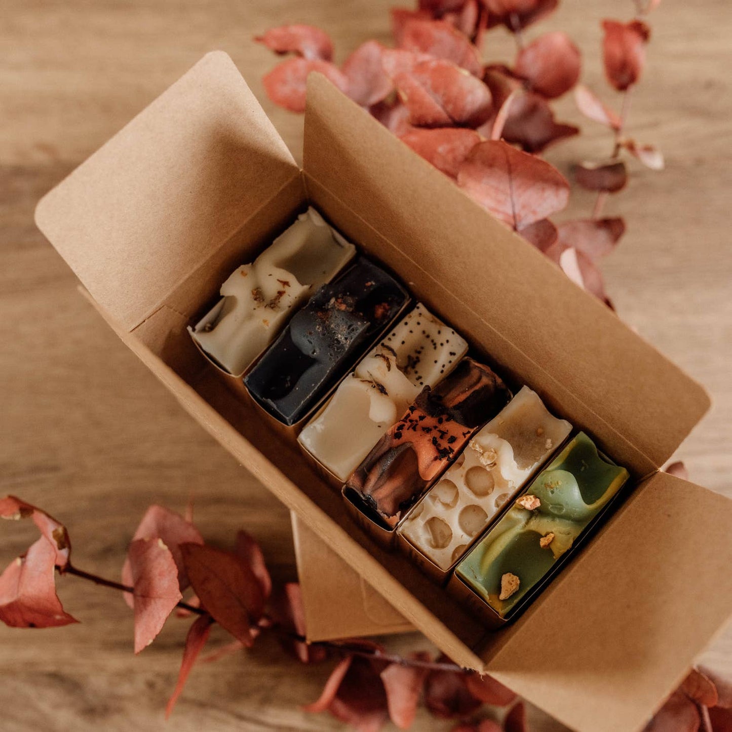 “Coffret kraft ouvert contenant six savons artisanaux Naturayl aux couleurs et textures variées, présenté sur une table en bois avec des feuilles séchées.”
