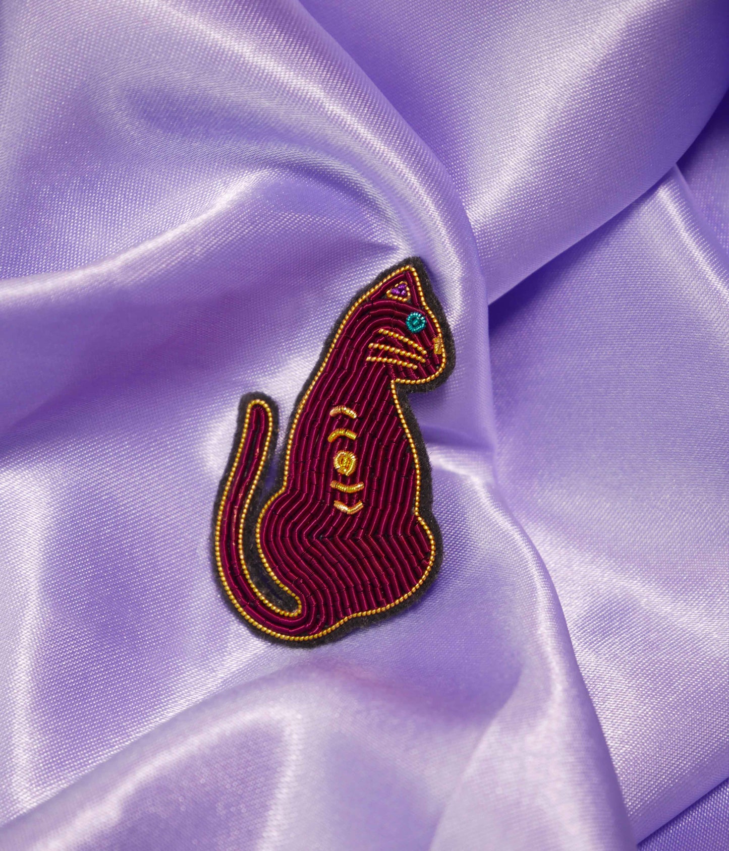 Broche Cat Magic Malicieuse, brodée à la main en fils dorés, posée sur tissu violet satiné