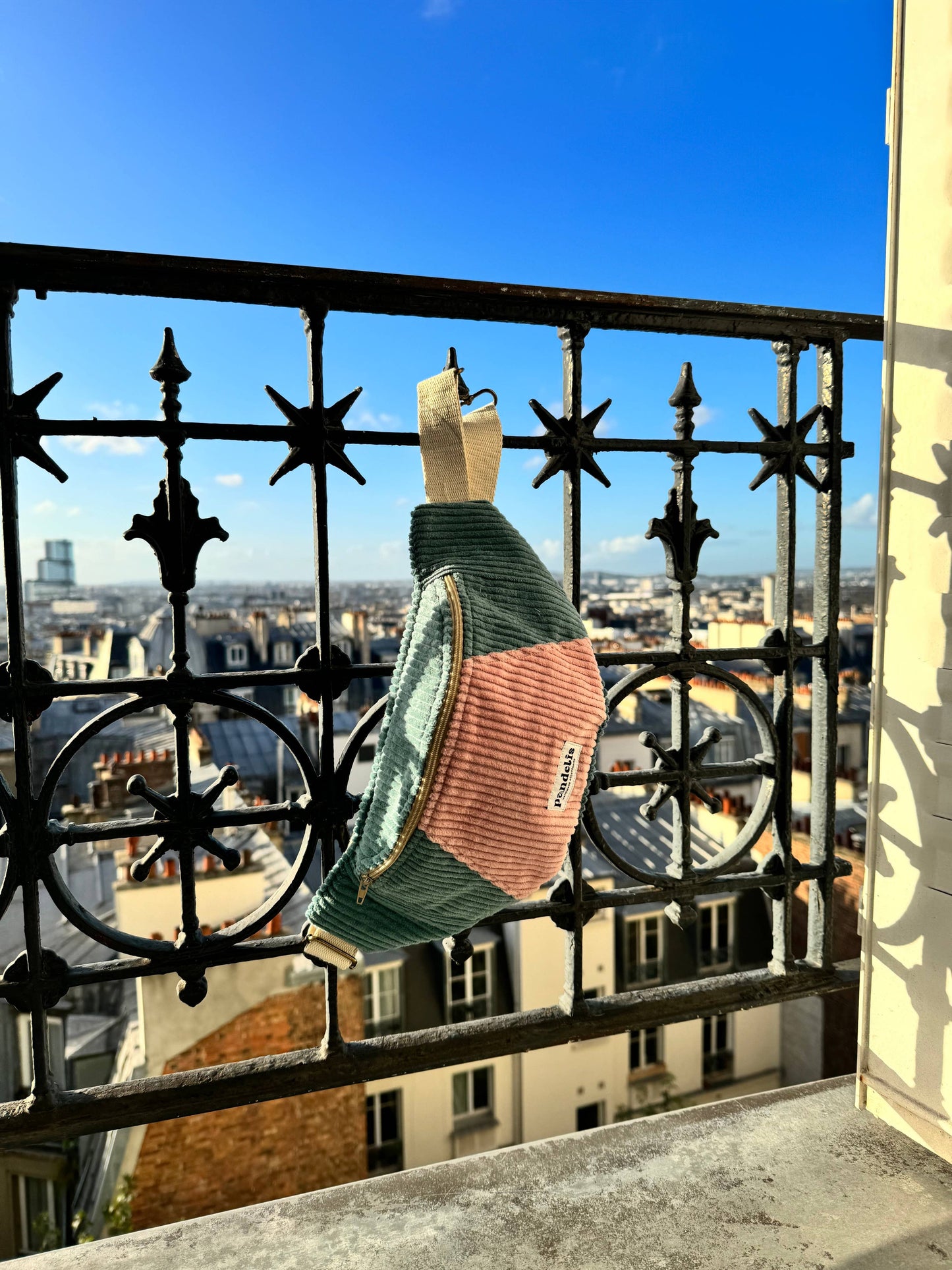 Banane en velours côtelé bicolore vert d’eau et rose Pandelis suspendue à un balcon parisien sous un ciel bleu, photographie lifestyle lumineuse.