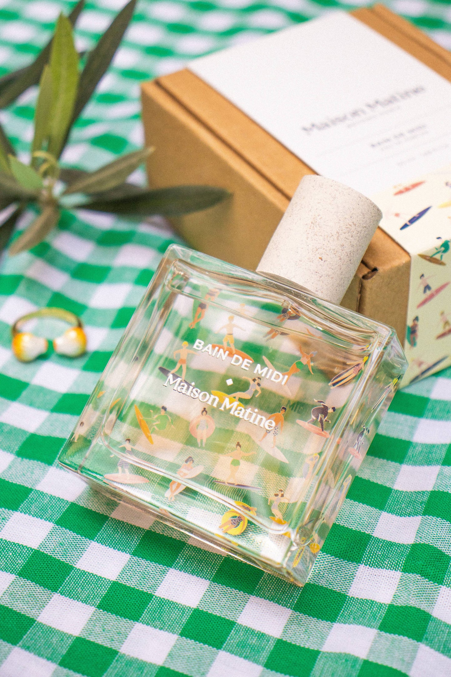 Flacon de parfum Bain de Midi Maison Matine sur une nappe à carreaux verts et blancs avec sa boîte kraft en arrière-plan.