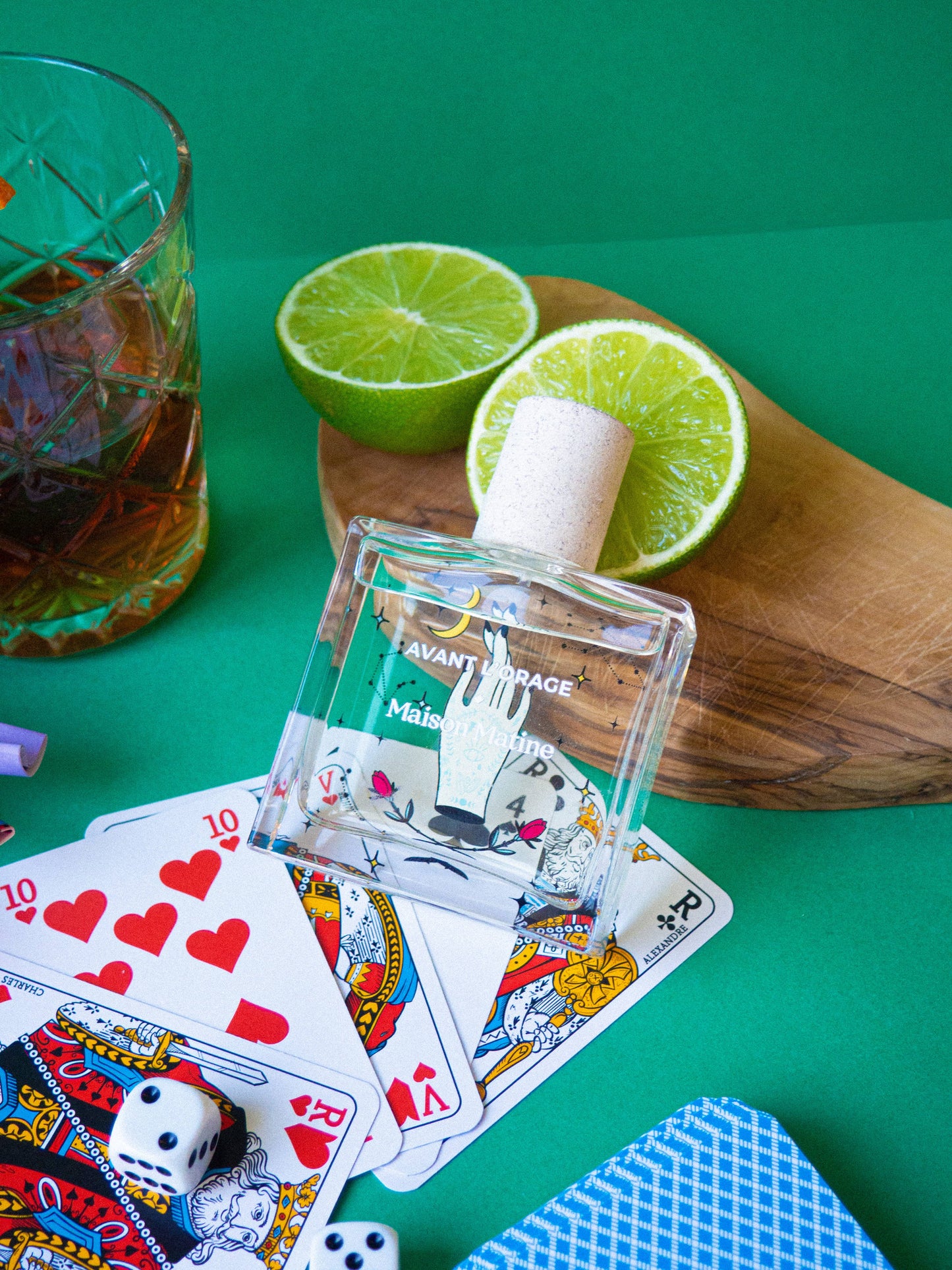 Flacon du parfum entouré de cartes à jouer et d’un cocktail lime-rhum sur fond vert.