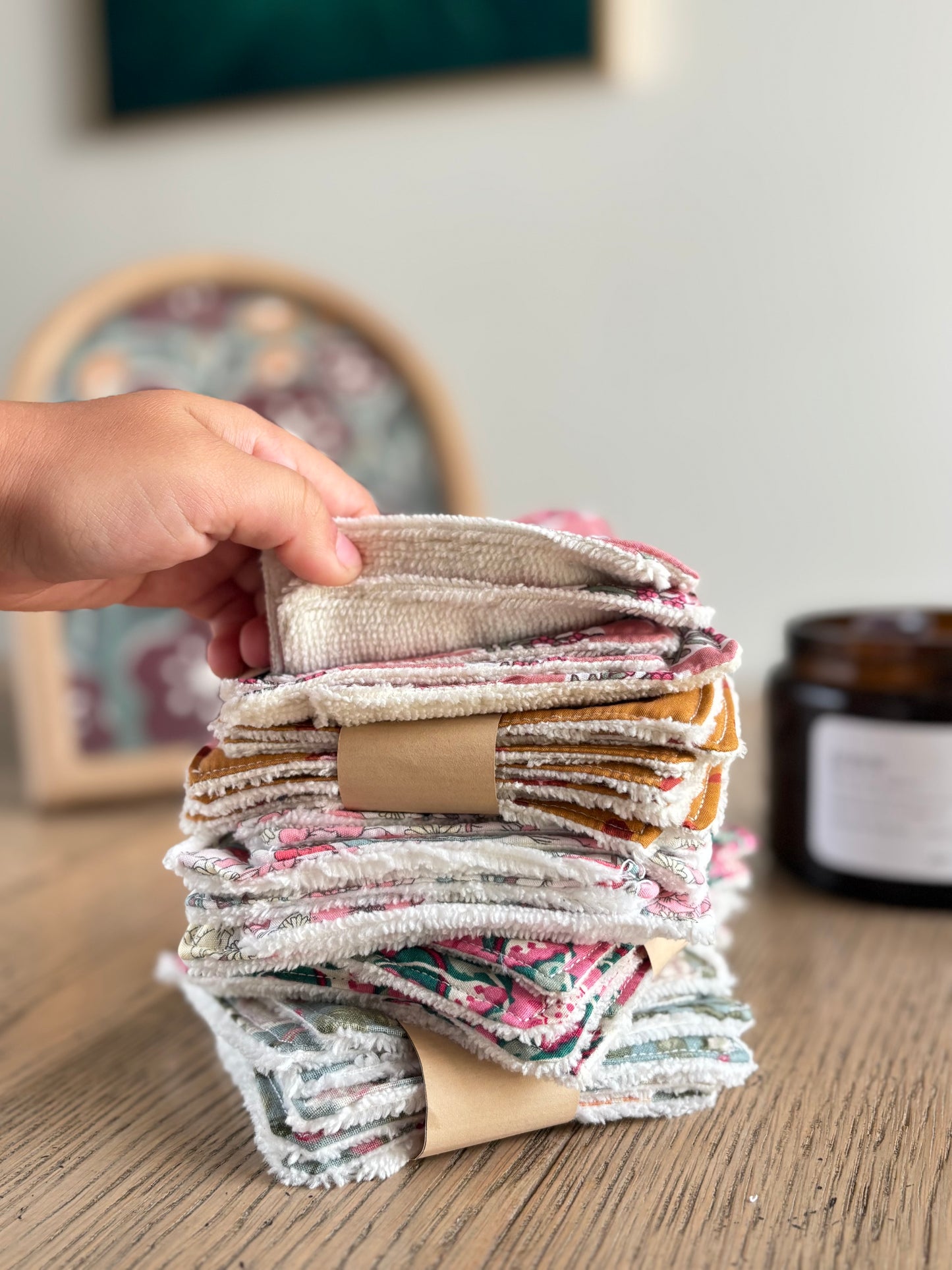 Main tenant une pile de lingettes lavables artisanales Marie en Août, aux motifs floraux variés, sur une table en bois naturel – accessoires zéro déchet Anahata.