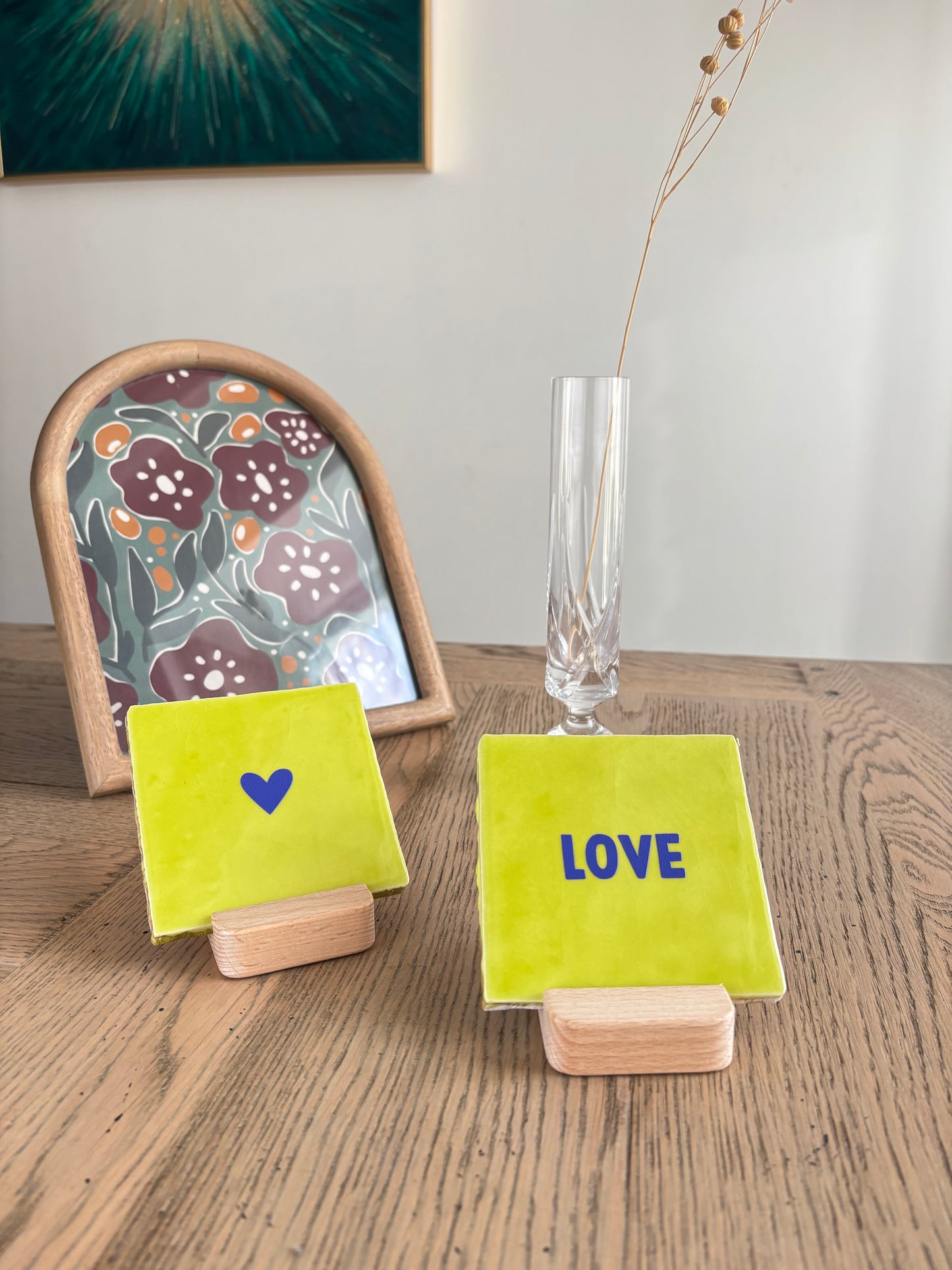 Carreaux décoratifs artisanaux Studio Meuk avec inscriptions LOVE et CŒUR bleus sur fond vert citron, posés sur supports en bois sur une table en bois clair. Création en céramique faite main disponible chez Anahata Concept Store.