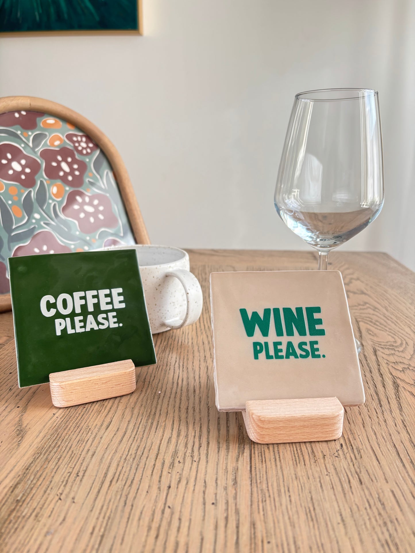 Carreaux décoratifs en céramique artisanale Studio Meuk, avec inscriptions “COFFEE PLEASE” vert forêt et “WINE PLEASE” vert sauge, posés sur supports en bois sur une table en bois clair. Créations faites main disponibles chez Anahata Concept Store.