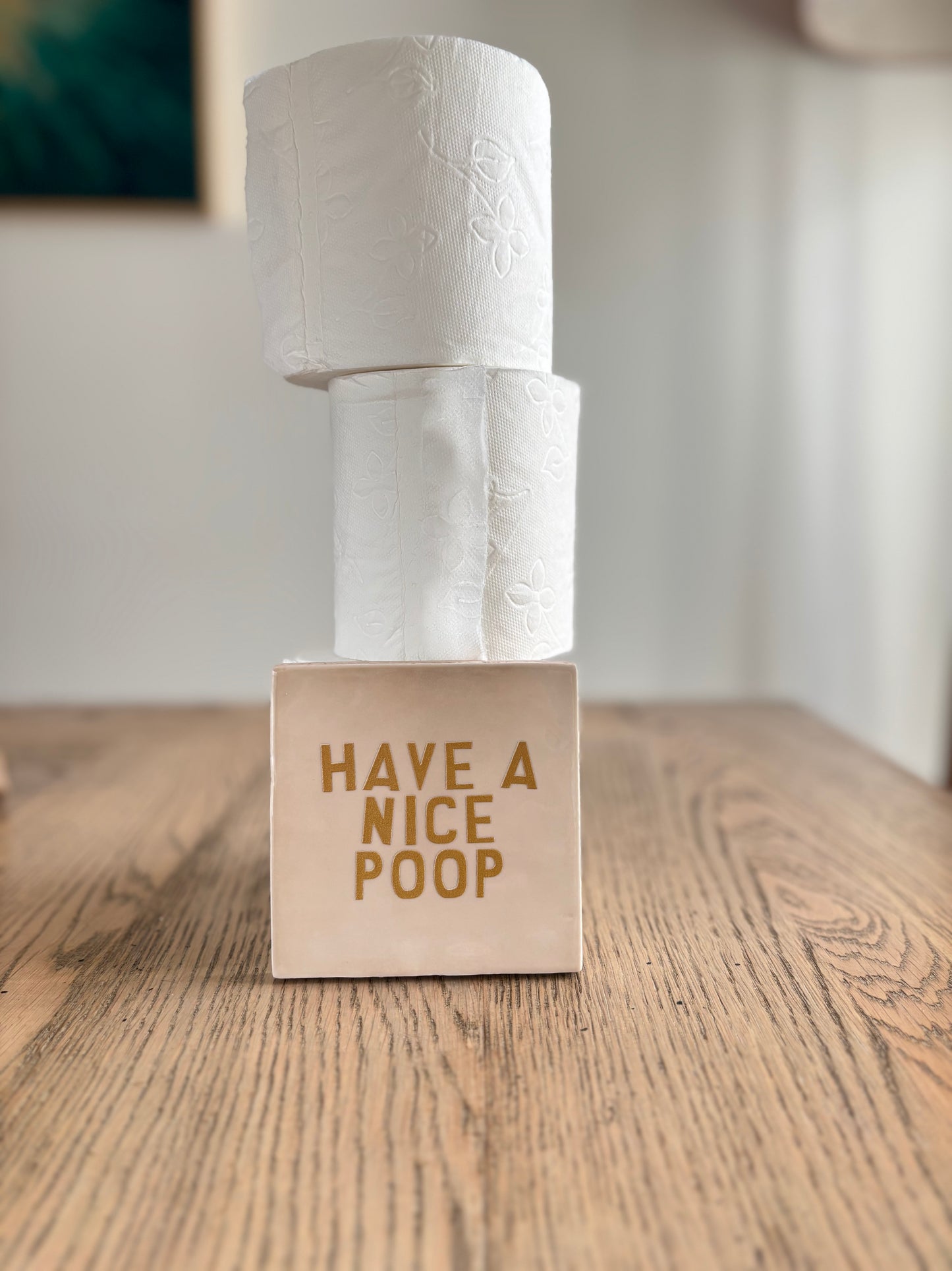 Carreau en céramique artisanale Studio Meuk avec inscription dorée “HAVE A NICE POOP”, accompagné de rouleaux de papier toilette empilés sur une table en bois. Décoration humoristique et faite main disponible chez Anahata Concept Store.