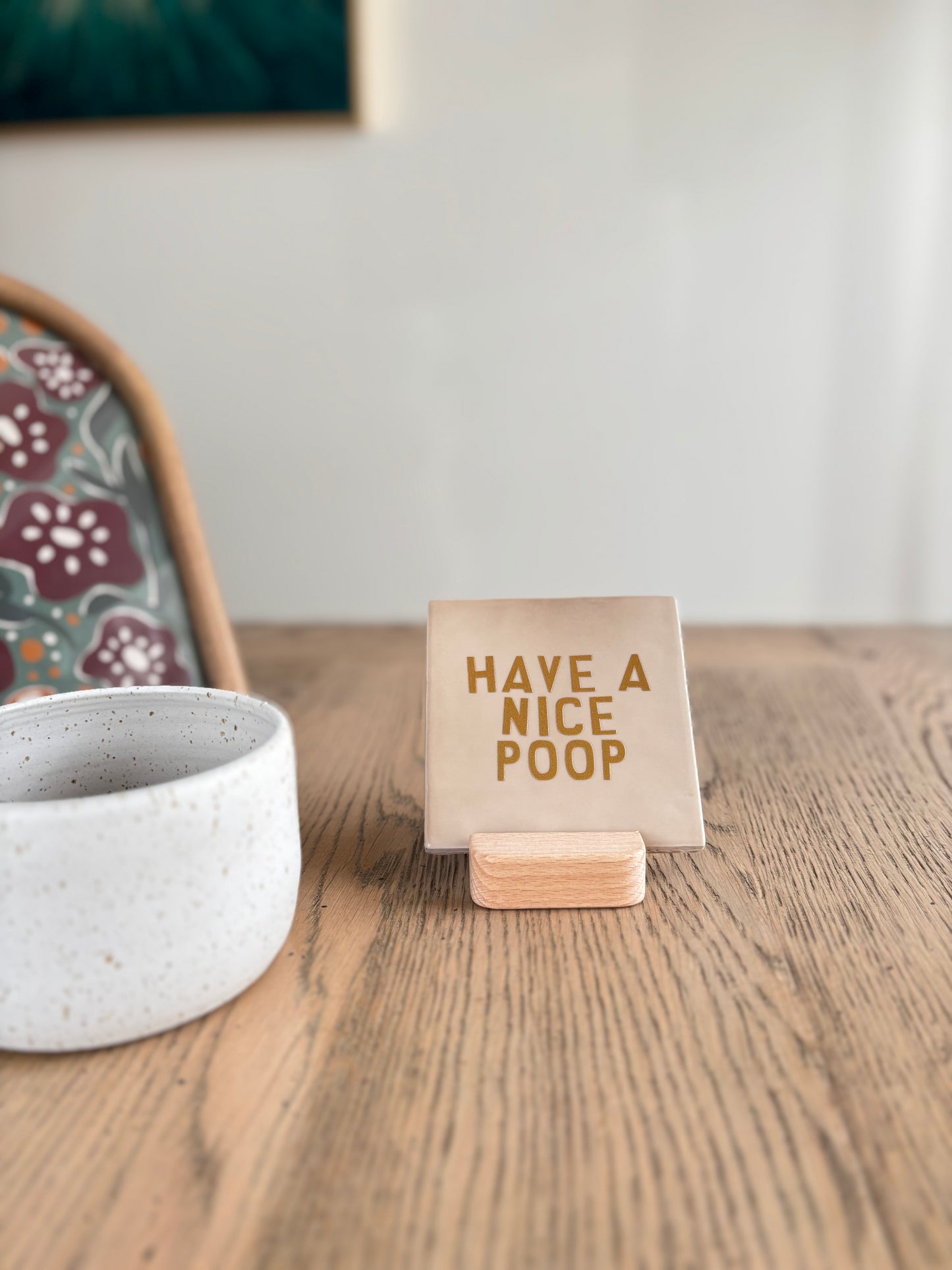 Carreau décoratif en céramique Studio Meuk avec message doré “HAVE A NICE POOP”, présenté sur un support en bois à côté d’une tasse blanche. Pièce artisanale à suspendre ou poser, vendue par Anahata Concept Store.