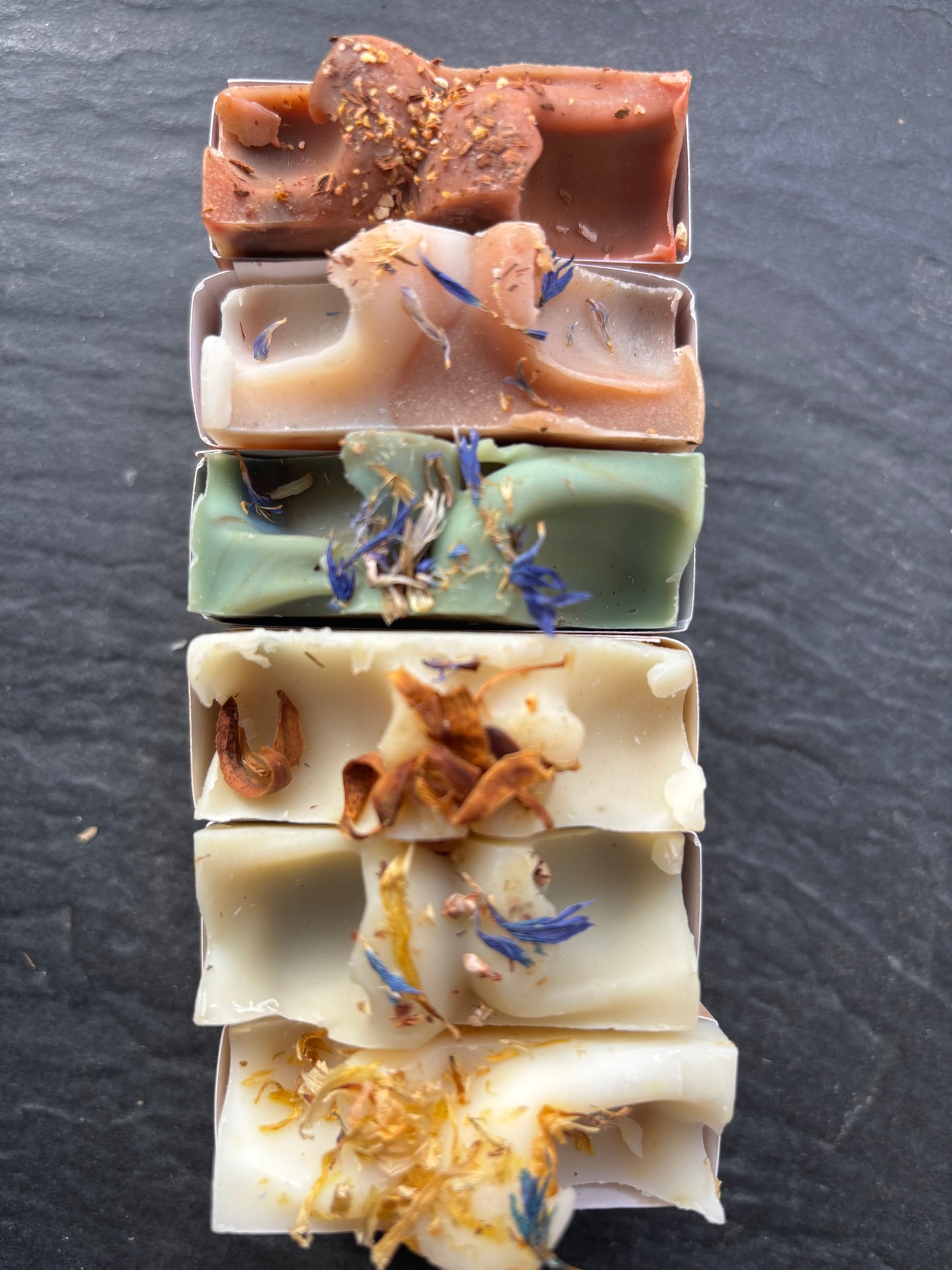 “Alignement de six savons artisanaux Naturayl coupés en morceaux, aux motifs végétaux et couleurs naturelles, posés sur une surface sombre.”