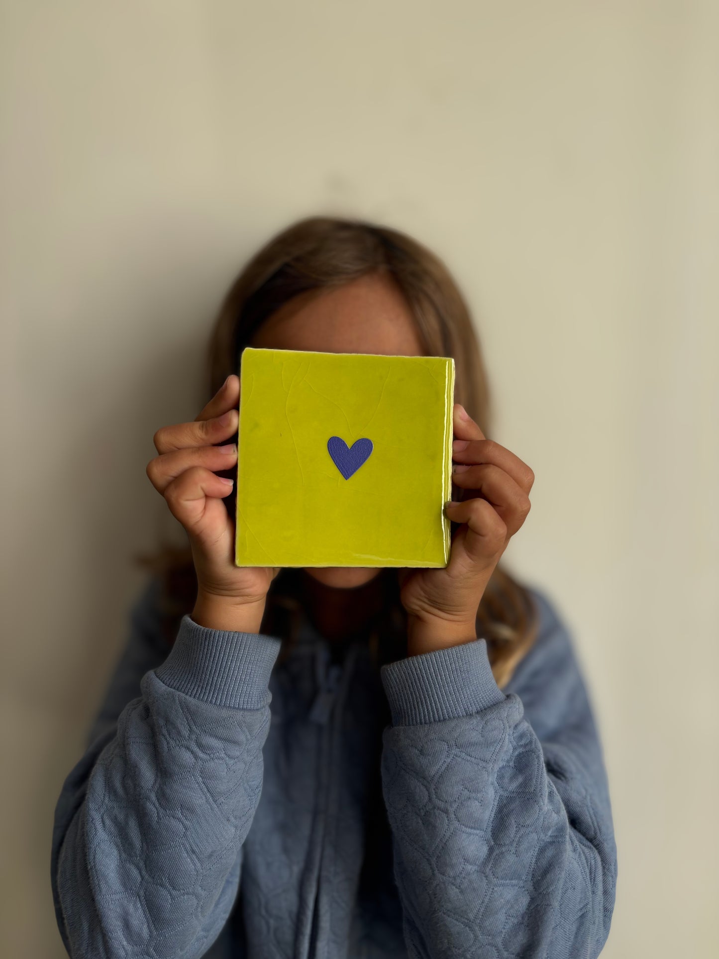 Carreau décoratif en céramique jaune citron avec un cœur bleu au centre, tenu entre les mains d’une personne en pull bleu. Création artisanale Studio Meuk, en vente sur Anahata Concept Store.