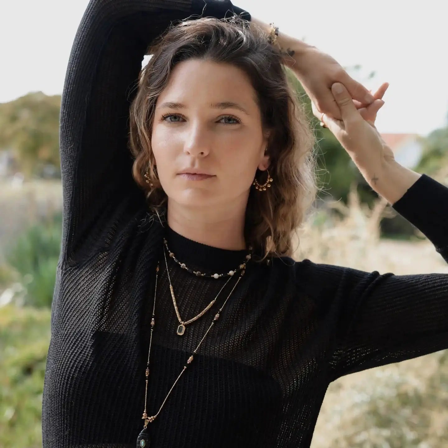 Femme portant le collier Clover et d’autres bijoux dorés, posant les bras levés en extérieur.