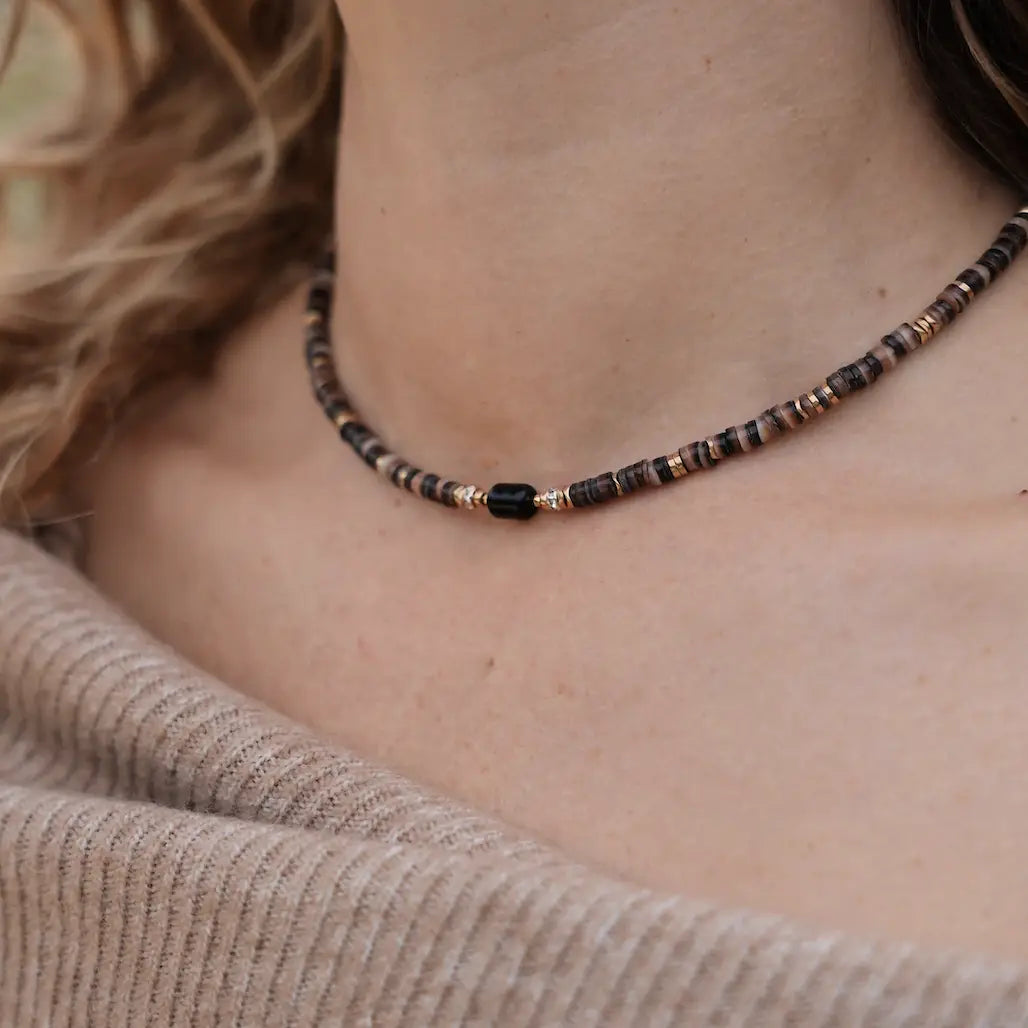 Gros plan du collier Lonnie porté, composé de perles Heishi marron et d’une pierre centrale noire, sur un décolleté en maille beige.