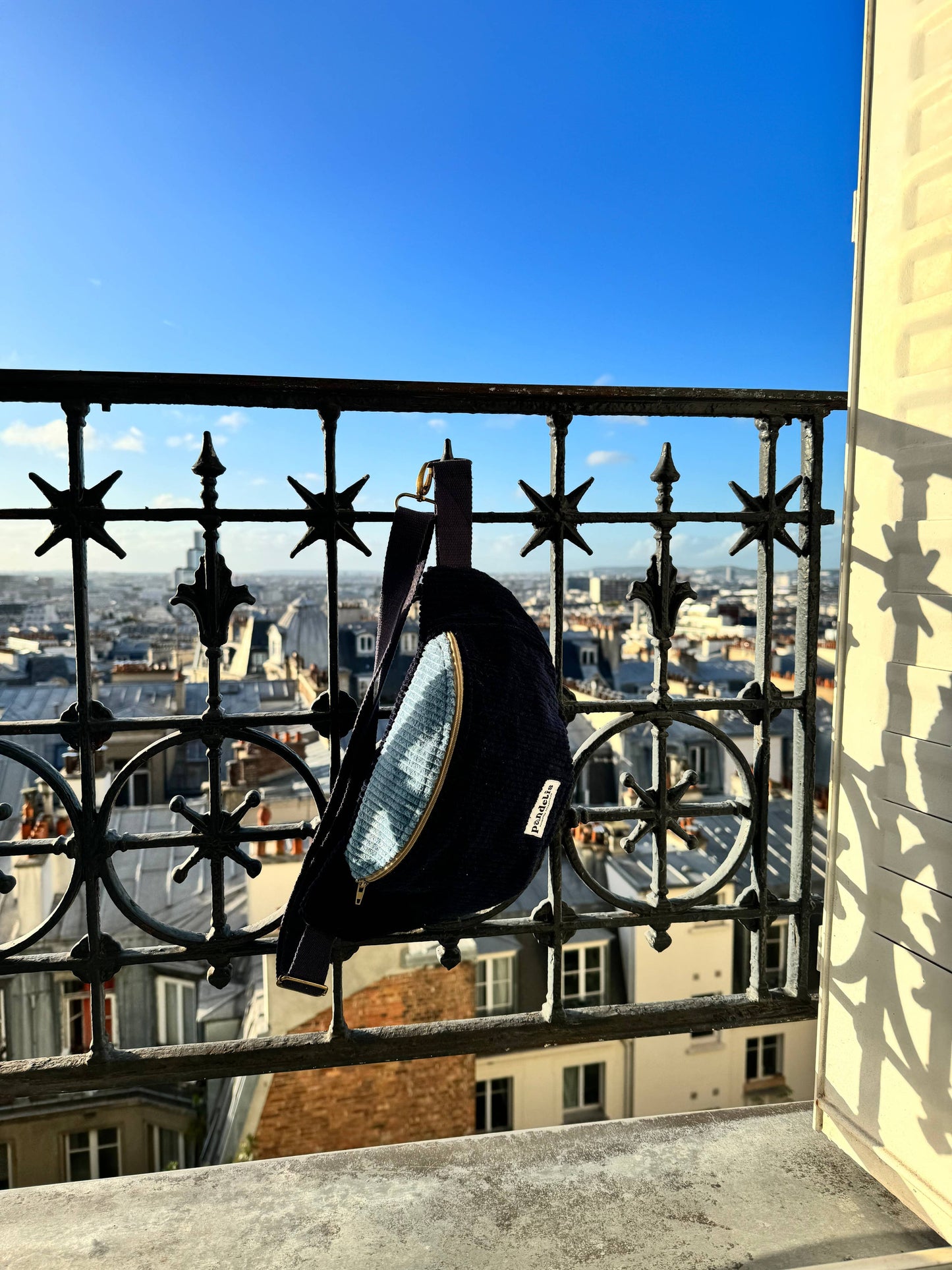 Banane bicolore en velours côtelé bleu marine et bleu ciel suspendue à une rambarde en fer forgé, photographiée avec une vue dégagée sur les toits de Paris sous un ciel bleu.