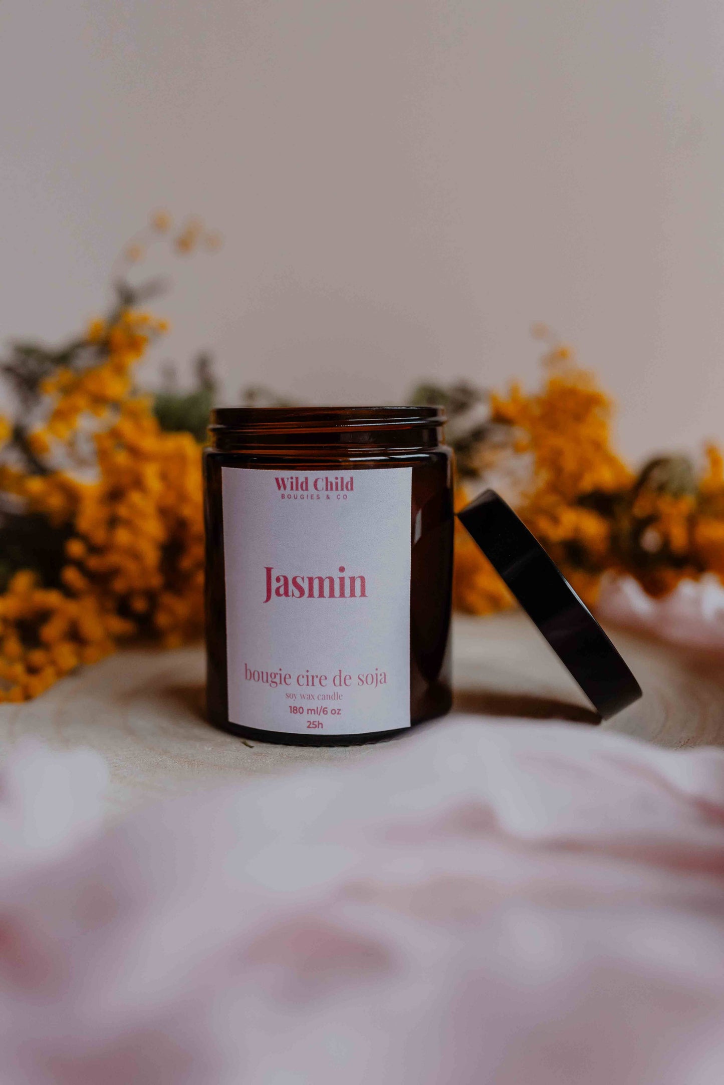 Bougie “Jasmin” en pot ambré, étiquette élégante, posée devant un fond doux décoré de fleurs jaunes séchées.