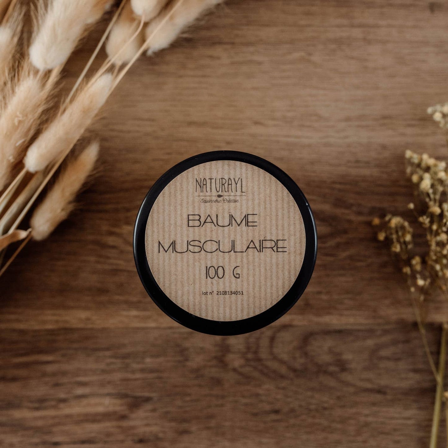 Vue du pot de baume fermé, étiquette visible (ambiance naturelle avec bois et fleurs séchées).