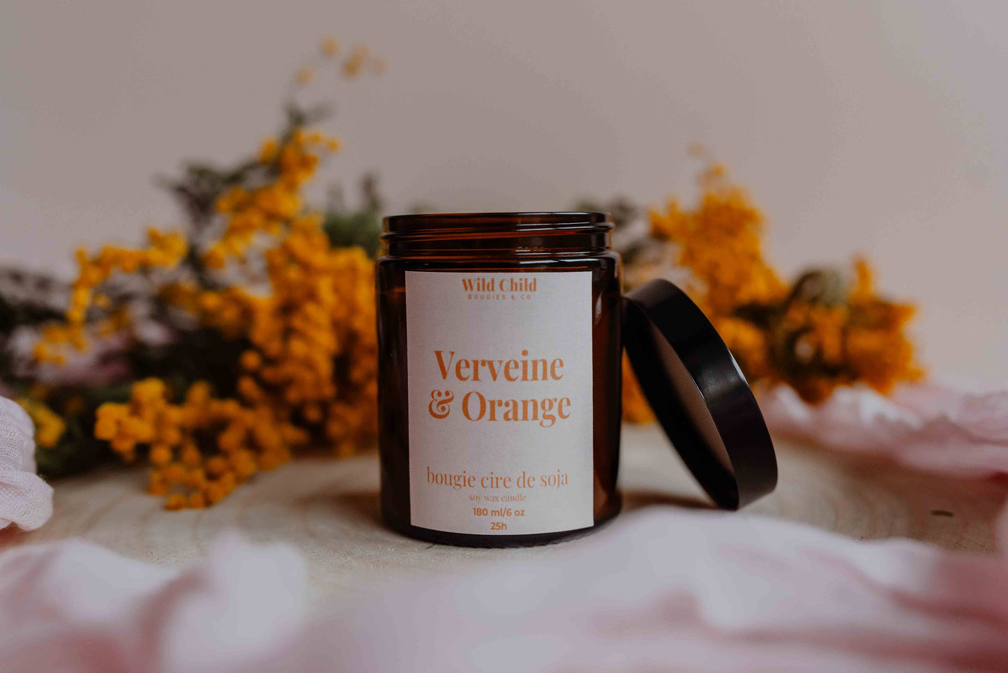 Bougie artisanale “Verveine & Orange” dans son pot ambré, posée sur un tissu doux avec bouquet de fleurs jaunes en arrière-plan.